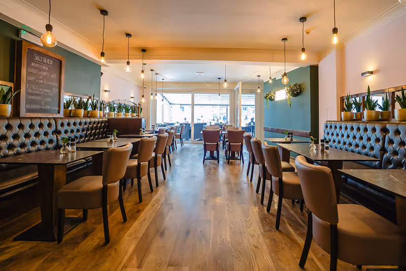 Modern restaurant interior with wooden floors, cushioned chairs, tufted bench seating, pendant lights, and potted plants along the walls.