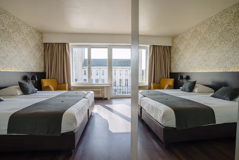 Modern hotel room with two beds, each with gray bed runners, pillows, and a bedside lamp, yellow armchairs, and a large window with beige curtains.