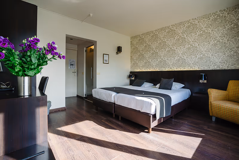 Bright hotel room with two single beds pushed together, dark wood floor, a yellow armchair, and a vase of purple flowers on a dark wood dresser.