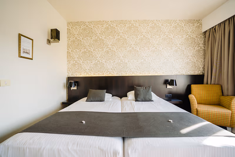 Minimalist hotel room with two single beds pushed together, each with a gray pillow and dark bed runner, a yellow armchair, patterned wallpaper, and brown curtains.