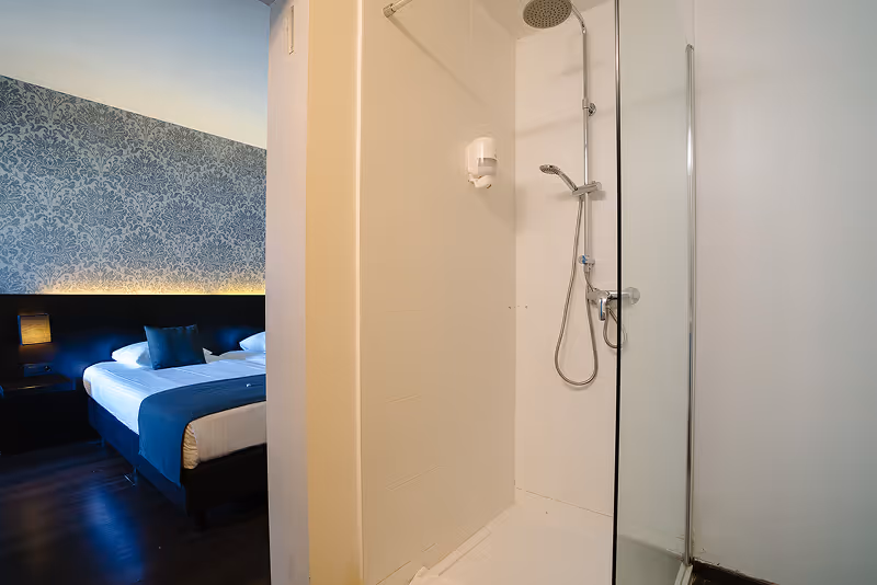 Modern bathroom shower area with rain showerhead and hand shower next to a bedroom with a bed and patterned wallpaper.