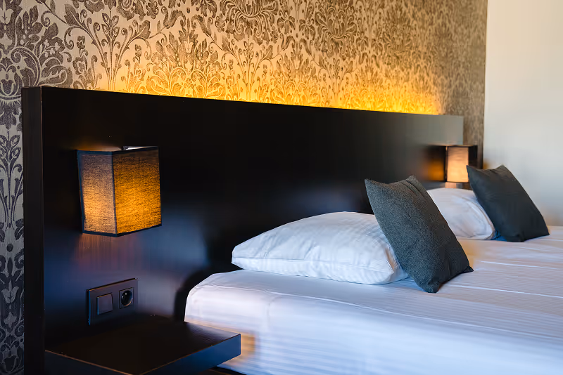 Modern hotel room with two single beds featuring white sheets, dark gray pillows, black headboard with built-in warm wall lights, and patterned wallpaper.