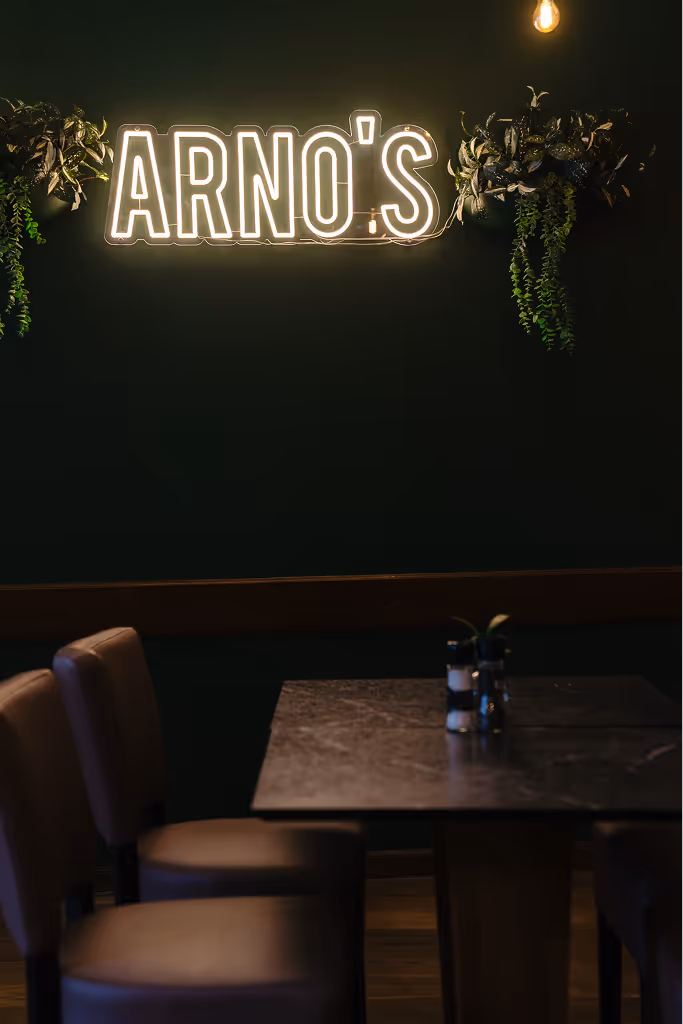 Dimly lit restaurant corner with a dark green wall, neon sign reading 'ARNO'S', hanging plants, and a table with four chairs.