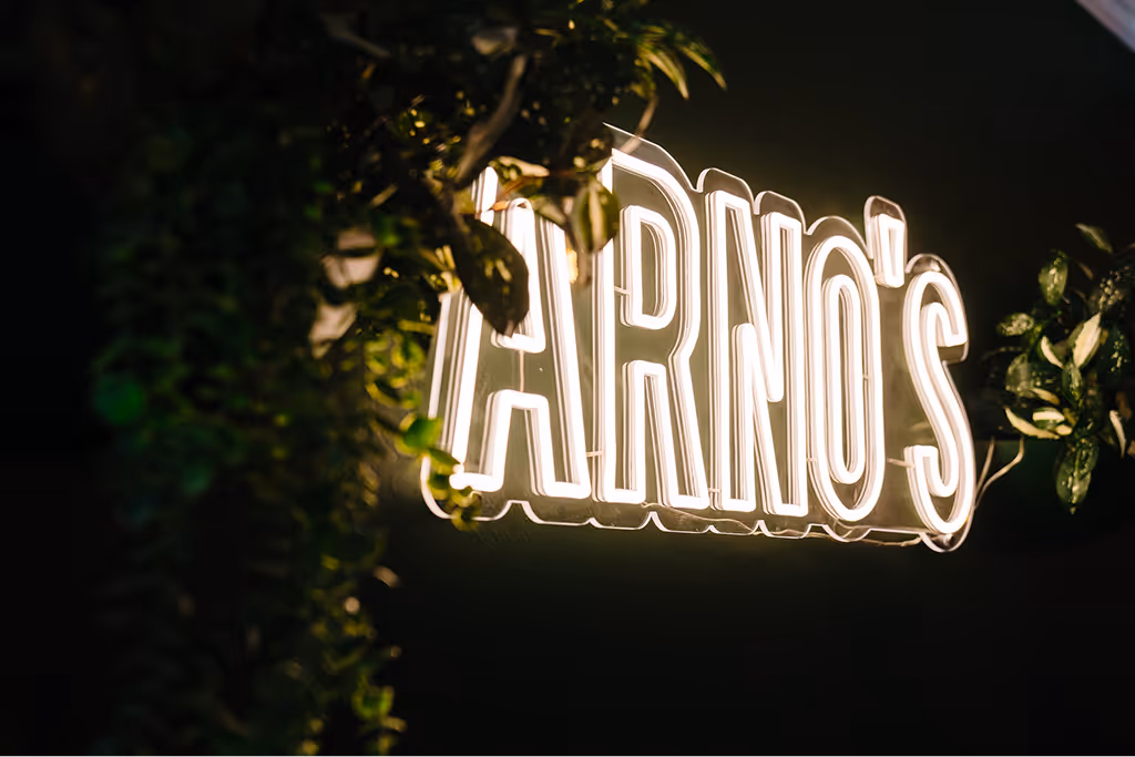 Illuminated sign with the text 'TARNO'S' surrounded by green foliage against a dark background.
