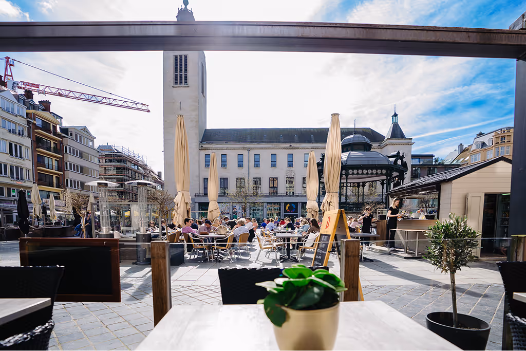 Outdoor café seating area with people dining under closed beige umbrellas in front of a large building with a tower and a gazebo on a sunny day.