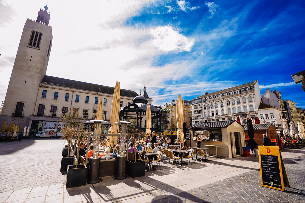 Outdoor café with people sitting at tables under closed umbrellas on a sunny day in a European city square with historic buildings and a tall tower in the background.