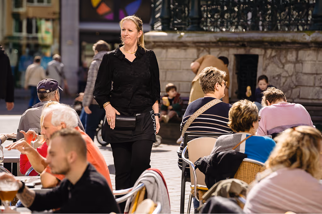 Woman wearing black walking through an outdoor café with people sitting and enjoying drinks under sunlight.