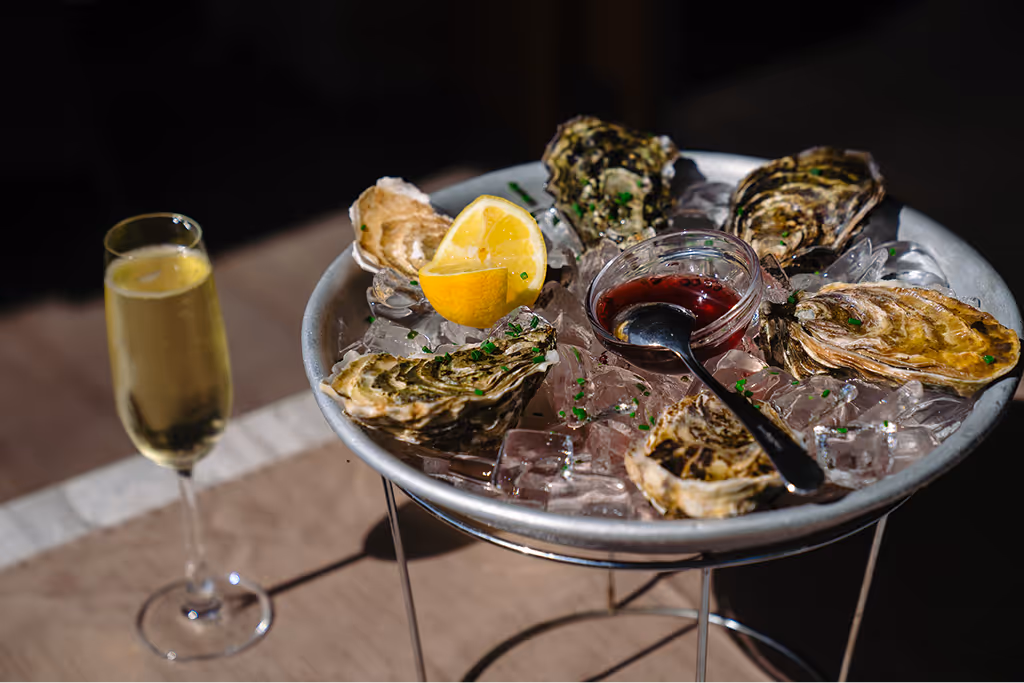 Plate of fresh oysters on ice with lemon wedge and red sauce, next to a glass of champagne on a table.