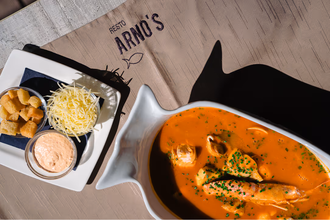 Seafood soup with shrimp, mussels, and herbs served in a fish-shaped bowl with side dishes of grated cheese, croutons, and sauce on a white plate.