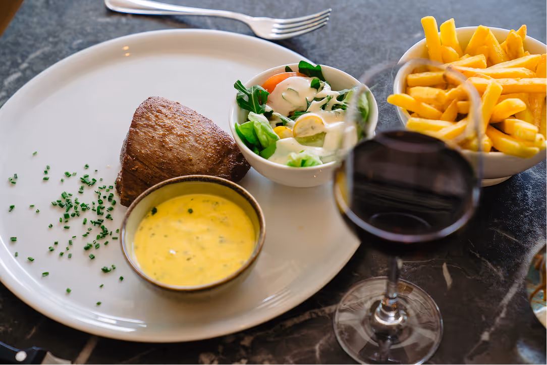 Plate with grilled steak, herb sauce, side salad, bowl of French fries, and a glass of red wine on a dark marble table.