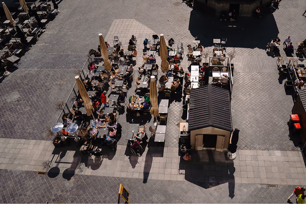Aerial view of people dining at outdoor café tables with umbrellas on a paved plaza.