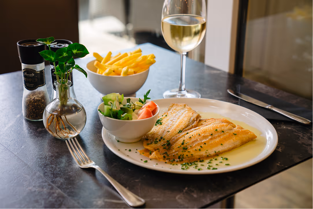 Plate with two cooked fish fillets garnished with chopped herbs, a bowl of salad, bowl of French fries, glass of white wine, and a small vase with green leaves on a black table.
