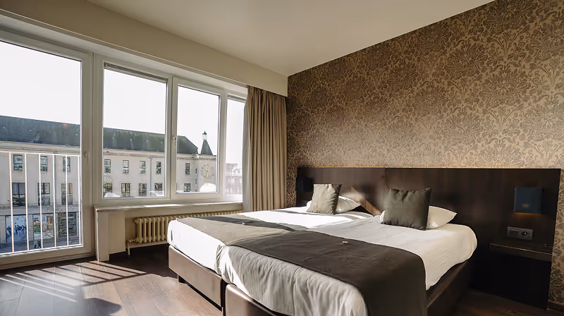 Sunlit hotel room with two single beds pushed together, dark headboard, patterned wallpaper, and large windows overlooking a street and buildings.