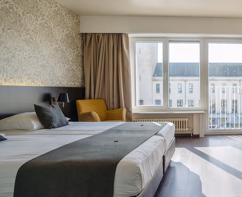Bright hotel room with a large bed, dark gray pillows, patterned wallpaper, a yellow armchair, and large windows letting in natural light.